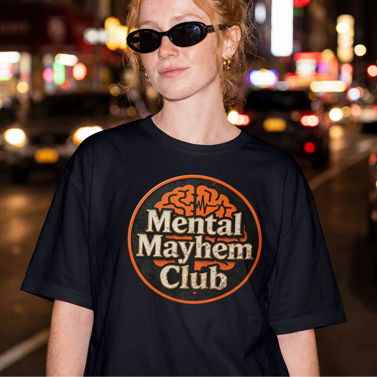 A woman wearing a black graphic t-shirt with a circular 'Mental Mayhem Club' logo, standing on a blurred city street at night.
