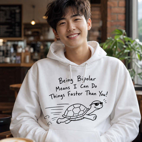 A smiling young man wearing a white hoodie with a black graphic print that reads 'Being Bipolar Means I Can Do Things Faster Than You!' and shows a cartoon turtle running. The photo is taken in a cozy, softly blurred cafe environment.