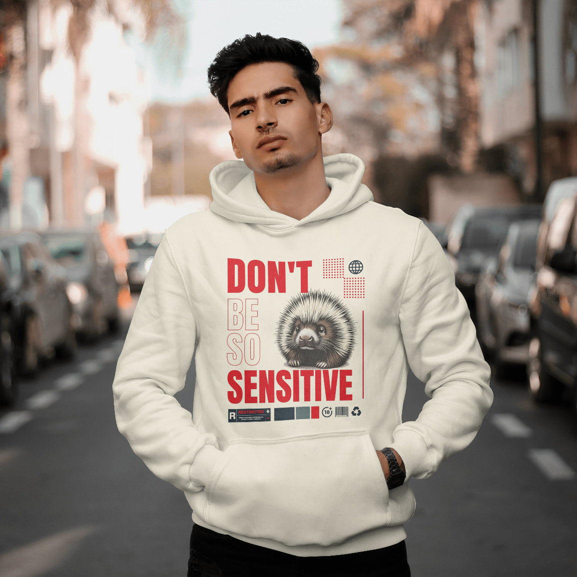 Man wearing a hoodie with a hedgehog graphic and text that reads "don't be so sensitive" on a city street.