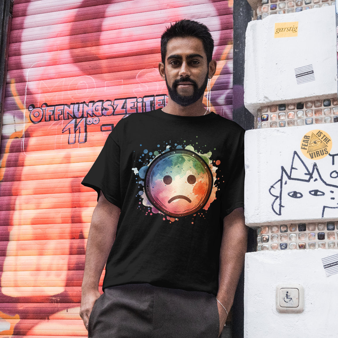 Man wearing a black t-shirt with a colorful sad face design, standing in front of a graffiti-covered wall.