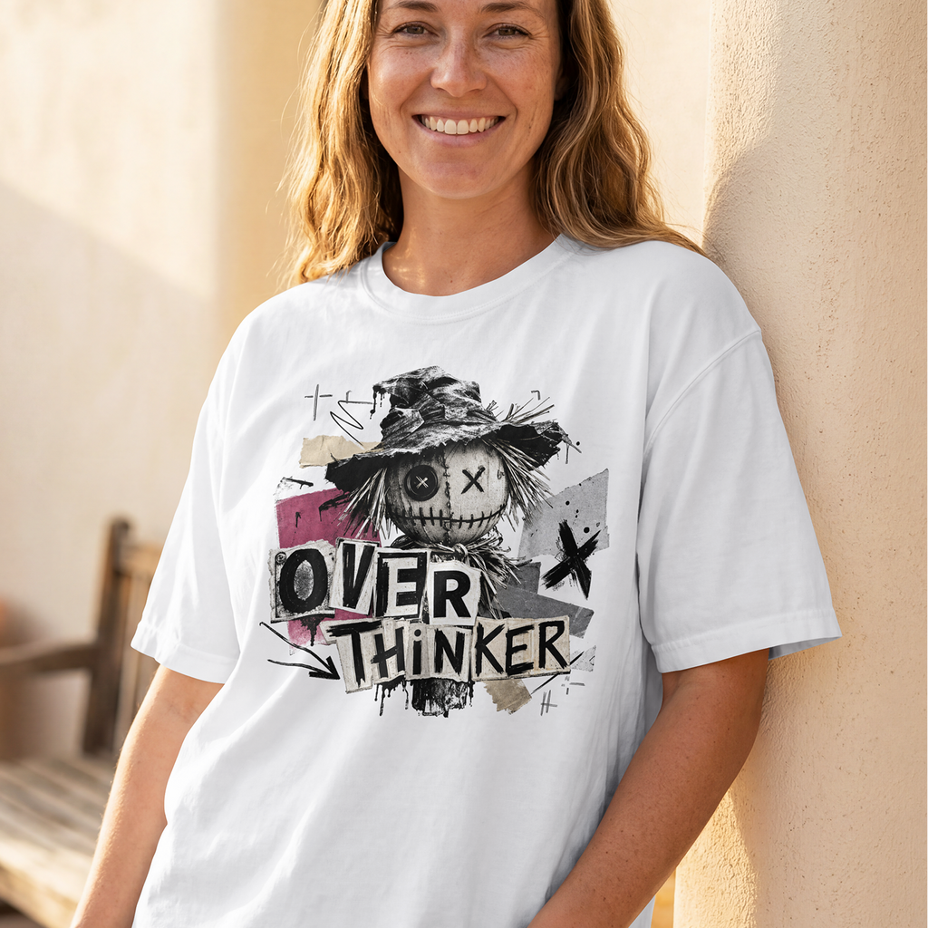 A smiling woman stands against a warm beige wall wearing a white graphic T-shirt. The shirt features a distressed black and gray illustration of a scarecrow with the words 'OVER THINKER' in bold lettering, with small pink and gray graphic accents.