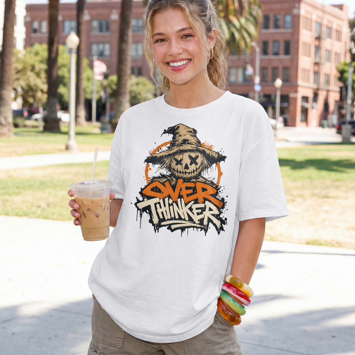 A smiling woman outdoors wearing a white graphic T-shirt with a distressed orange, black, and tan 'Over Thinker' skull-and-hat design. She holds an iced coffee in a park-like setting with buildings and trees in the background.