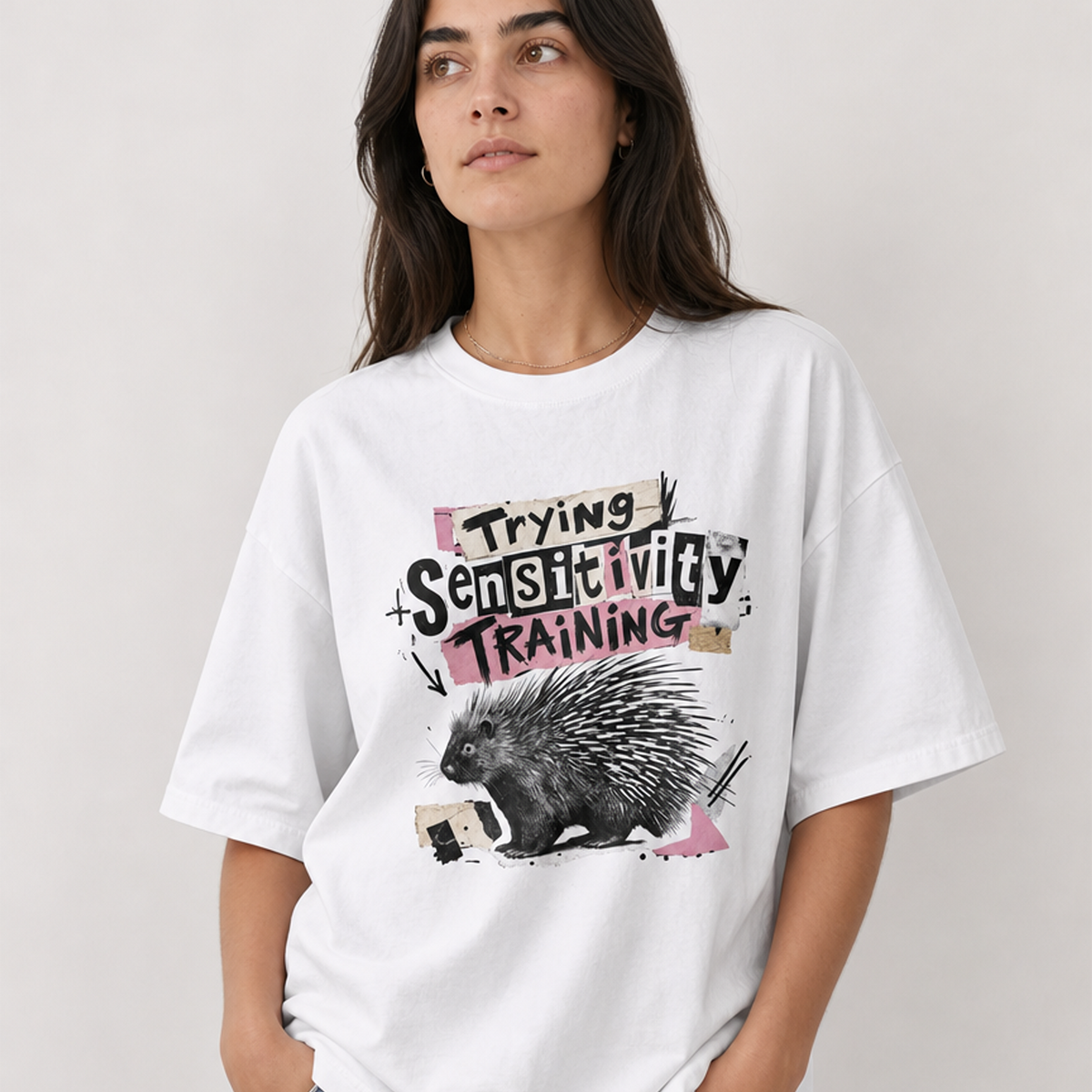 A woman wearing a white oversized graphic T-shirt with a porcupine illustration and the text 'Trying Sensitivity Training' in pink, black, and beige tones, paired with light blue jeans against a light gray studio background.