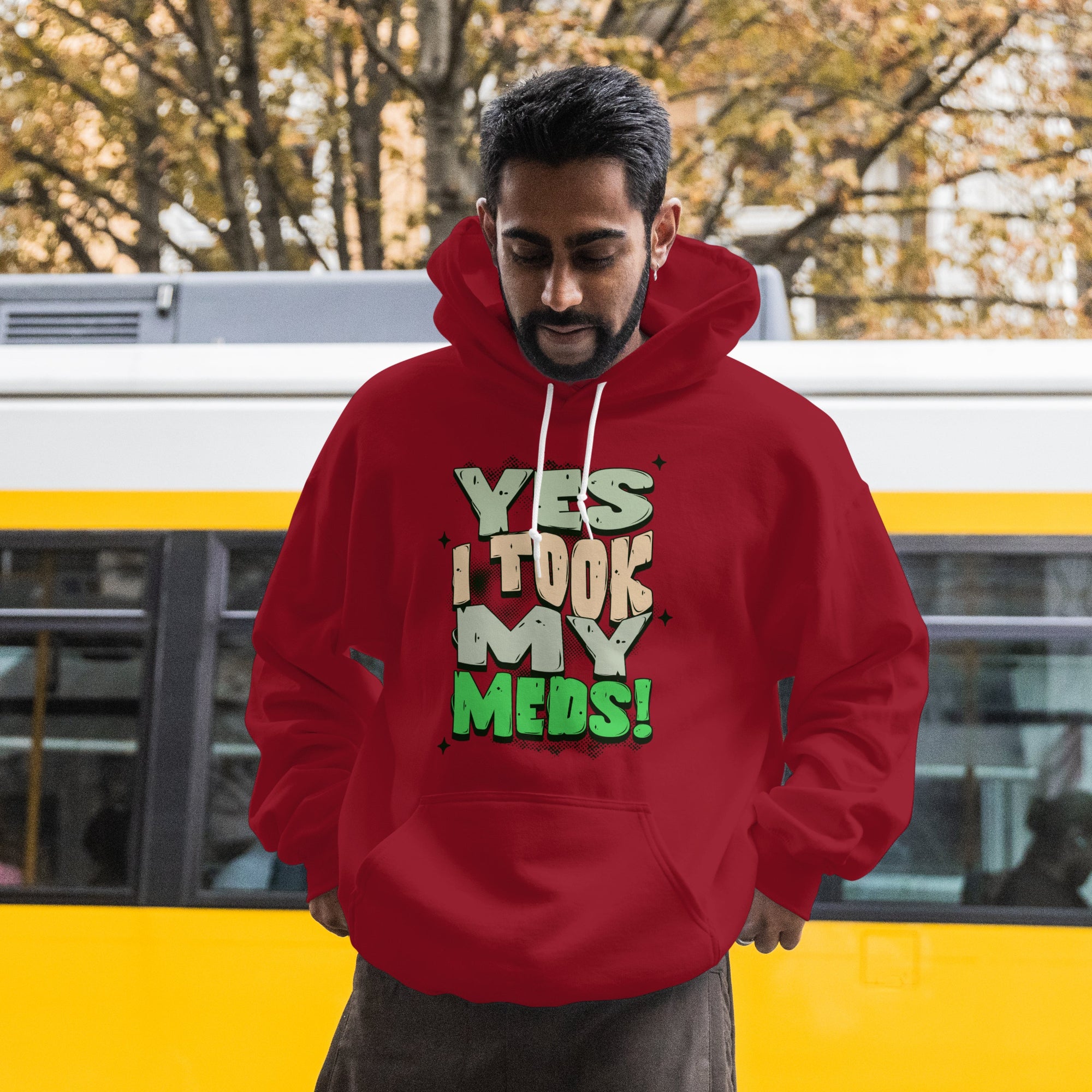 Man wearing a red hoodie with text that reads, yes, I took my meds, standing in front of a yellow bus.